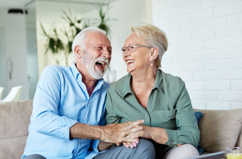 Independent living for couples as two older adults sit together on a sofa, laughing and holding hands during a relaxed moment in a bright, comfortable home.
