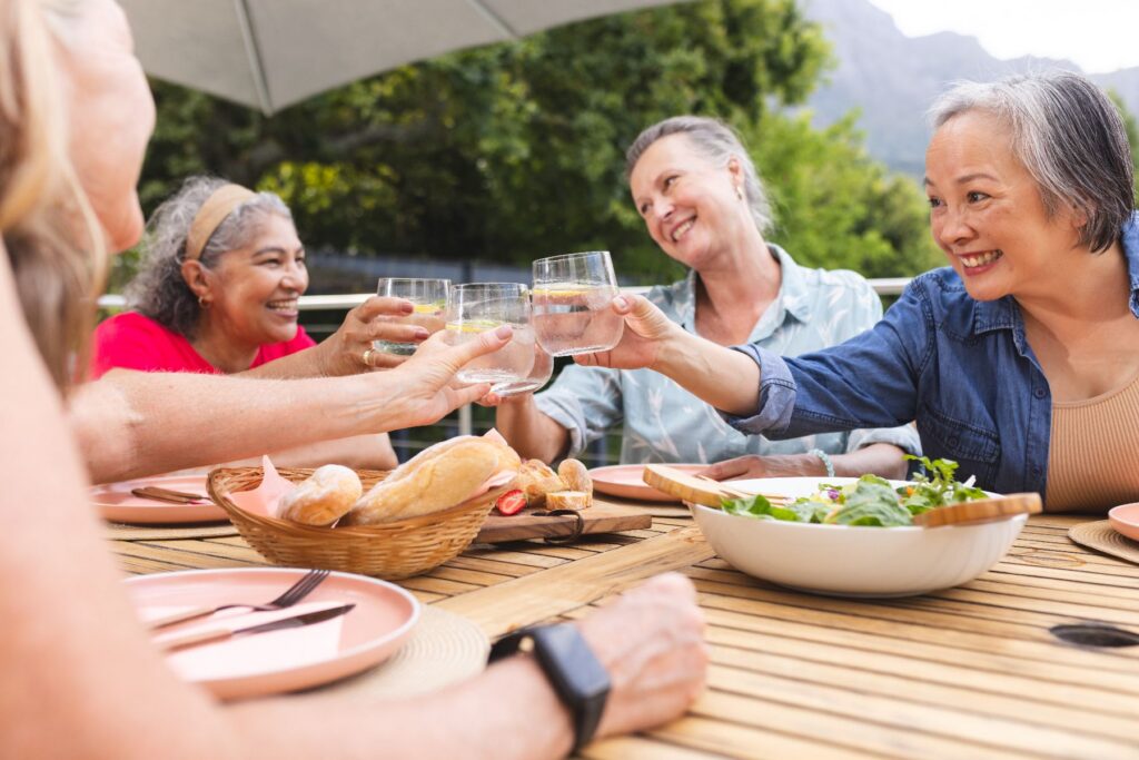A group of senior women raising glasses in a toast at an outdoor table, enjoying the social connection and senior routine benefits of sharing a sunny afternoon meal together.