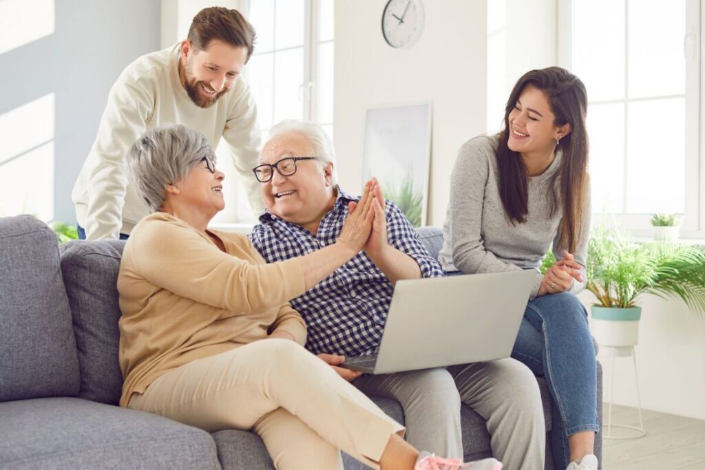 An older couple sits together on a couch sharing a joyful high-five while an adult son leans in from behind and a daughter looks on smiling, a laptop open on the couple's lap — a warm family moment that speaks to the kind of connection families weigh when comparing senior living vs. staying at home.