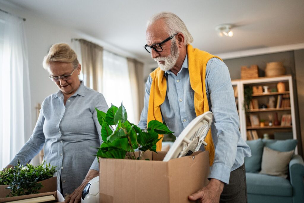 An older couple packing belongings in a bright living room, symbolizing the decision-making process between moving to a senior living apartment and staying at home.